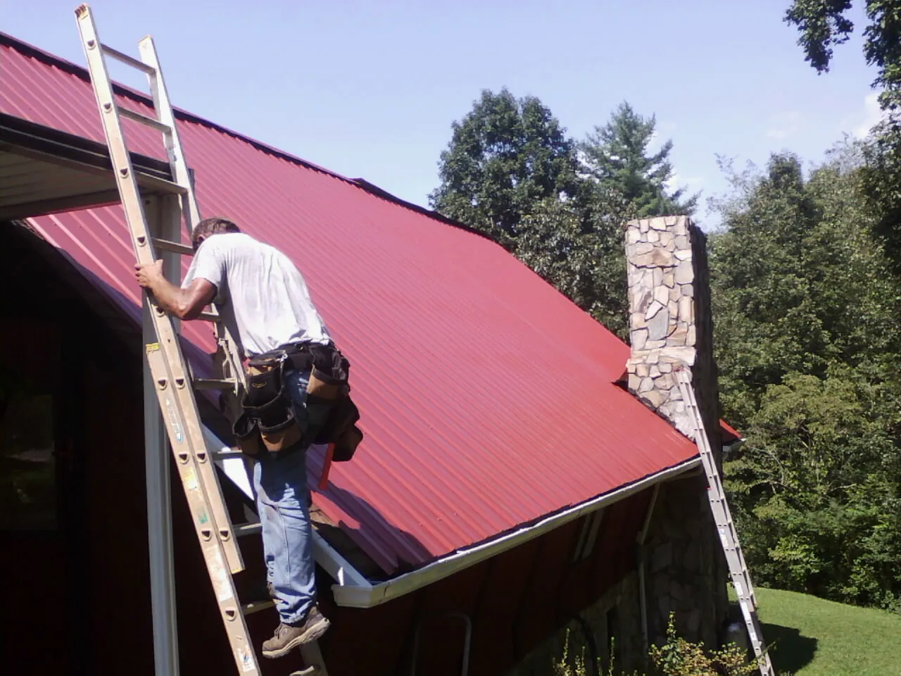 Roofer on a ladder beside a red metal roof and stone chimney — Soffit & Fascia Repair by Roofing Brothers in Des Plaines